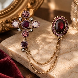 May include: A gold-toned brooch featuring a cross-shaped component adorned with pink, purple, and iridescent crystals, connected by two delicate gold chains to a separate oval-shaped piece centred with a large, deep red faceted crystal.