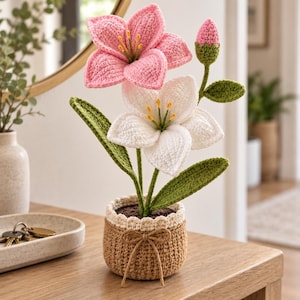 Può includere: Una pianta in vaso all'uncinetto con un giglio rosa, un giglio bianco e un piccolo bocciolo rosa. I fiori hanno stami gialli e steli verdi con foglie. La pianta è contenuta in un vaso all'uncinetto color marrone chiaro con bordo crema e un fiocchetto.