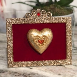 Puede incluir: Un adorno dorado en forma de corazón con una joya roja en el centro, enmarcado en un marco dorado adornado con un fondo de terciopelo rojo.