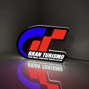 May include: A glowing LED sign featuring the Gran Turismo logo with a blue and red stylised GT emblem. The text reads Gran Turismo The Real Driving Simulator in white lettering against a black background, with a power cord extending from the side.