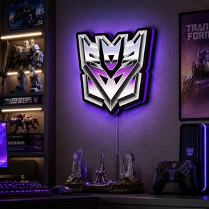 Può includere: Un'insegna da parete con il logo Decepticon caratterizzata da una finitura argento metallizzato e retroilluminazione a LED viola. L'emblema presenta un design angolare netto con accenti neri, creando un effetto luminoso sulla parete.