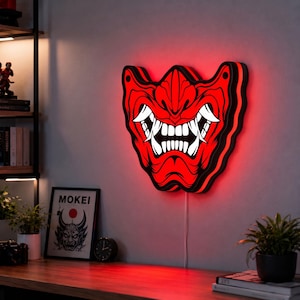 May include: A wall-mounted LED sign shaped like a red and black Hannya mask with prominent white fangs and a fierce expression. The sign emits a bright red glow from behind its edges, creating a vibrant silhouette against the wall surface.
