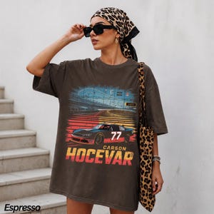May include: An espresso-colored oversized t-shirt featuring a graphic print of a race car with the number 77, a racetrack background, and the text Carson Hocevar in bold yellow and red lettering. The shirt has a crew neckline and short sleeves.