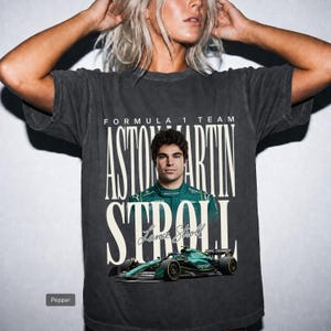 May include: A charcoal grey short-sleeve t-shirt featuring a graphic print of Formula 1 driver Lance Stroll in a green racing suit. The text reads Formula 1 Team, Aston Martin, and Stroll, with a green Aston Martin race car at the bottom.