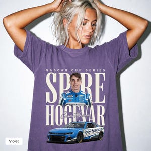 May include: A violet short-sleeve t-shirt featuring a graphic print with the text NASCAR CUP SERIES, SPIRE, HOCEVAR, and Carson Hocevar. The design includes a portrait of a race car driver and a blue number 77 Spectrum race car at the bottom.