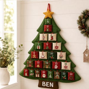 May include: A green felt Christmas tree wall hanging advent calendar with twenty-four numbered pockets. Each pocket features festive holiday icons like Santa, reindeer, and candy canes. A yellow star sits at the top and the name BEN is at the base.