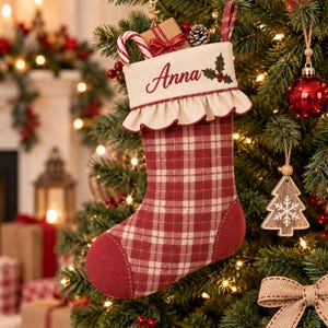 May include: A red and cream plaid Christmas stocking featuring a cream-coloured cuff with a ruffled edge, embroidered with the name Anna in red script and a holly sprig. The stocking has a solid red toe and heel section and a hanging loop.