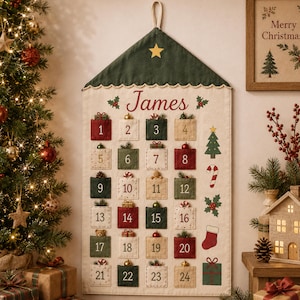 May include: A fabric wall-hanging advent calendar with a cream-colored background, featuring a dark green roof peak with a yellow star. It has twenty-four numbered pockets in red, green, and cream, personalized with the name James in red script.