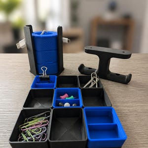 Pode incluir: Um conjunto organizador de secretária modular com componentes de plástico preto e azul. Inclui um suporte vertical para bobina de cabo, um suporte preto em forma de H e uma grelha de nove recipientes de armazenamento quadrados e retangulares.
