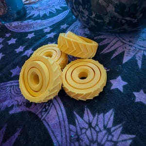 May include: A collection of four bright yellow, circular, 3D-printed objects featuring a distinct chevron tread pattern along the outer edge and a concentric, stepped circular design on the face, each with a central hole for mounting or assembly.