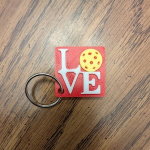 May include: A square red keychain featuring the white text LOVE, where the letter O is replaced by a yellow pickleball with small circular indentations. A silver metal split ring is attached to the bottom left corner of the red square base.