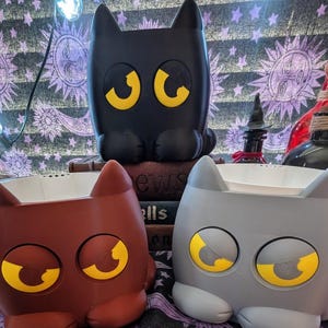 May include: Three cat-shaped planters in black, brown, and gray. Each planter features large, stylized yellow eyes and a white interior basin. The black cat planter is positioned behind the brown and gray planters, which sit side by side in the front.