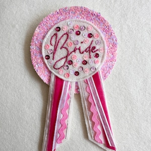 May include: A circular badge featuring a pink glitter border and a white centre embroidered with the word Bride in dark pink script. The centre is decorated with scattered pink and white sequins. Two ribbons with pink and white trim hang below.