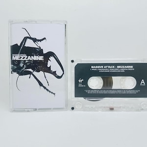May include: A clear plastic cassette case with a white insert featuring a black beetle graphic and the text Massive Attack Mezzanine, alongside a clear cassette tape with a black label listing tracks including Angel, Risingson, and Tear Drop.