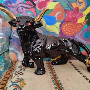 May include: A glossy black ceramic bull figurine with gold-colored horns and accents. The bull has red eyes and is posed in a charging stance. The figurine is on a patterned surface, with a glass head and colorful artwork in the background.