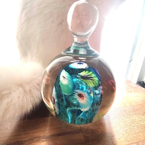 May include: A clear glass perfume bottle with a round body and a clear stopper. The interior features a colorful design with blue, green, and white floral elements. The bottle sits on a wooden surface.