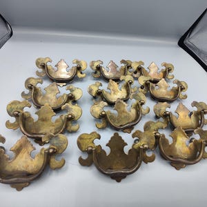 May include: A collection of ten brass drawer pulls featuring a decorative backplate with ornate, curved edges and a swinging bail handle. Each piece displays a weathered, aged patina with varying tones of gold and brown across the metal surface.
