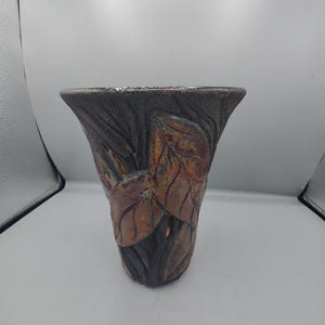 May include: A flared ceramic vase featuring a dark, textured charcoal-colored base with raised, embossed leaf motifs in shades of copper, bronze, and iridescent metallic tones. The vessel has a wide, open rim and a tapered body with a sculptural finish.