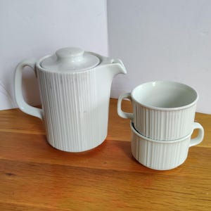 May include: White ceramic tea set with a ribbed design. The set includes a teapot with a lid and two stacked teacups with handles. The teapot has a spout and a handle. The set is on a wooden surface.