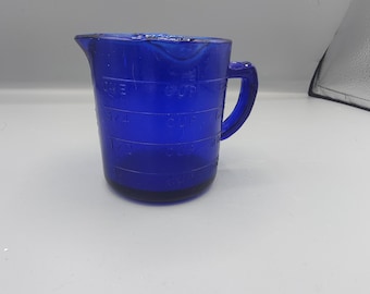 May include: A cobalt blue glass measuring cup featuring a handle and a pouring spout. The exterior surface is embossed with measurement markings in cups, including 1/4, 1/2, 3/4, and 1 cup increments, clearly visible against the translucent blue glass.