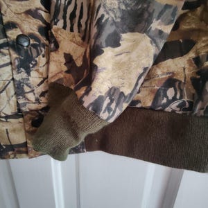 K&ouml;nnte beinhalten: Tarnjacke mit Knopfverschluss und olivgr&uuml;nen Rippb&uuml;ndchen und Saum. Die Jacke hat ein Muster aus braunen, beigen und schwarzen Bl&auml;ttern und Zweigen.