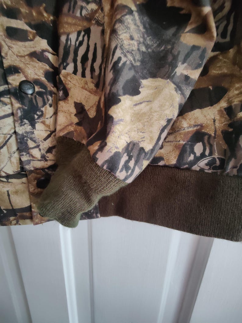 K&ouml;nnte beinhalten: Tarnjacke mit Knopfverschluss und olivgr&uuml;nen Rippb&uuml;ndchen und Saum. Die Jacke hat ein Muster aus braunen, beigen und schwarzen Bl&auml;ttern und Zweigen.