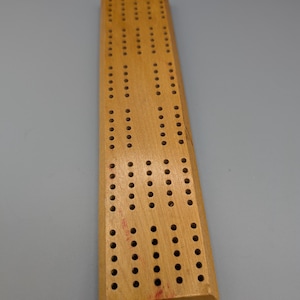 May include: A rectangular light brown wooden cribbage board featuring a series of drilled holes arranged in parallel tracks for scorekeeping. The board has a smooth, natural wood grain finish with visible signs of wear and small red markings.