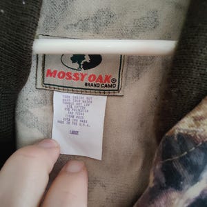 K&ouml;nnte beinhalten: Nahaufnahme eines Camouflage-Stoffs mit einem Etikett "Mossy Oak Brand Camo". Das Etikett enth&auml;lt Waschanweisungen und die Gr&ouml;&szlig;e "LARGE". Der Stoff ist eine Mischung aus Baumwolle und Polyester.