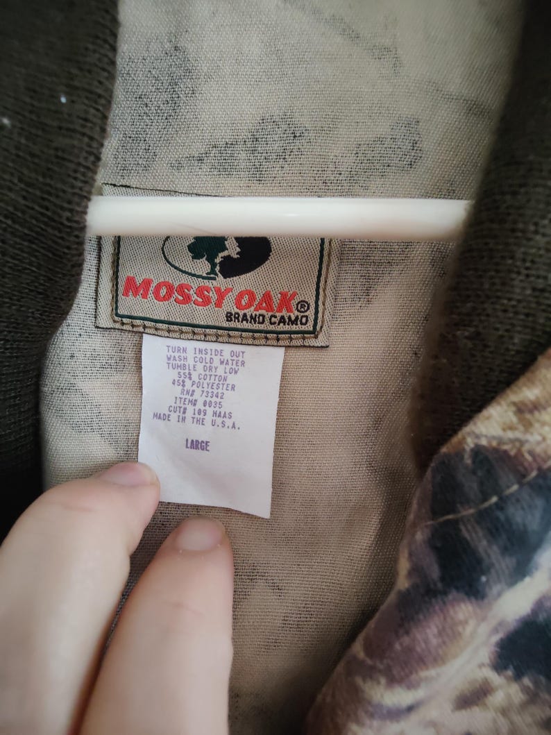 K&ouml;nnte beinhalten: Nahaufnahme eines Camouflage-Stoffs mit einem Etikett "Mossy Oak Brand Camo". Das Etikett enth&auml;lt Waschanweisungen und die Gr&ouml;&szlig;e "LARGE". Der Stoff ist eine Mischung aus Baumwolle und Polyester.