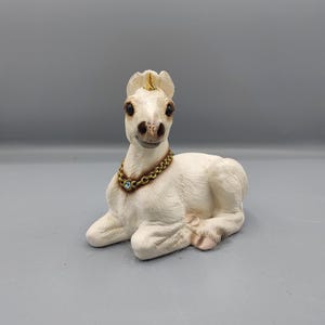 May include: A cream-colored unicorn figurine in a reclining position. The creature features a small gold-colored horn on its forehead, dark eyes, and a textured coat. It wears a gold-toned chain necklace adorned with a single light blue stone.