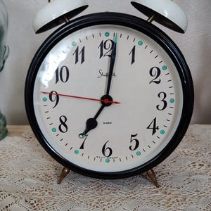 May include: A vintage Sheffield alarm clock with a black frame, white face, and gold-colored accents. The clock features black numerals, a red second hand, and two white bells on top. The words "Sheffield" and "West Germany" are printed on the face.