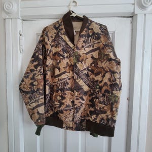 K&ouml;nnte beinhalten: Eine Camouflage-Bomberjacke mit braunem Kragen, B&uuml;ndchen und Bund. Die Jacke hat ein Muster aus braunen, beigen und gr&uuml;nen Bl&auml;ttern und &Auml;sten. Sie h&auml;ngt an einem wei&szlig;en Kleiderb&uuml;gel vor einer wei&szlig;en T&uuml;r.