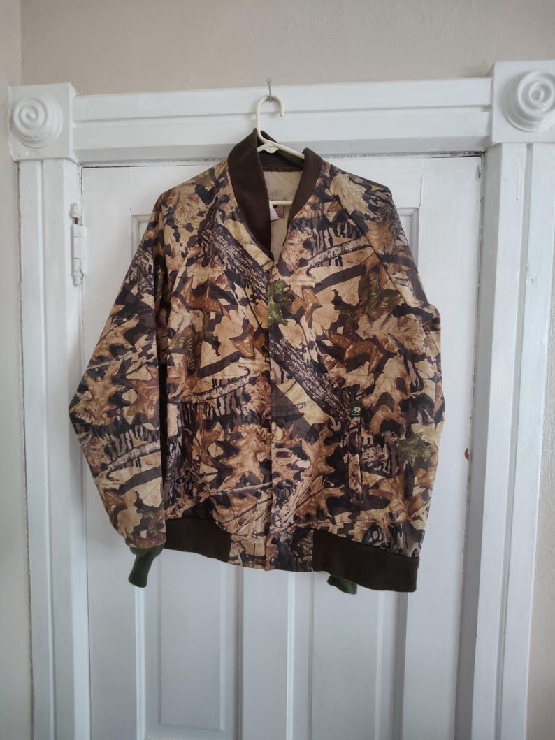 K&ouml;nnte beinhalten: Eine Camouflage-Bomberjacke mit braunem Kragen, B&uuml;ndchen und Bund. Die Jacke hat ein Muster aus braunen, beigen und gr&uuml;nen Bl&auml;ttern und &Auml;sten. Sie h&auml;ngt an einem wei&szlig;en Kleiderb&uuml;gel vor einer wei&szlig;en T&uuml;r.