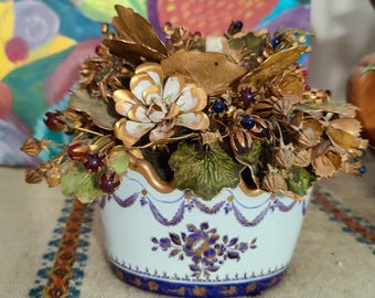 May include: A decorative ceramic vase with a floral arrangement. The vase is white with blue floral patterns and gold trim. The arrangement features gold-colored leaves and flowers, with accents of green and red. The overall theme is vintage and decorative.