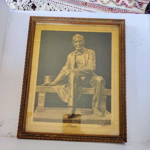 May include: A framed black and white print of a bronze statue of Abraham Lincoln sitting on a bench. The print is in a gold-colored frame with a decorative border. The print is signed "A. Lincoln" in the bottom right corner.