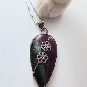 May include: A silver necklace with a teardrop-shaped pendant. The pendant is a mix of purple, green, and black hues, with two small silver flower accents. The chain is a delicate silver link.