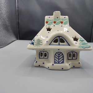 May include: A cream-colored ceramic house-shaped candle holder featuring blue polka dots, painted green pine trees, and brown accents. The structure includes cut-out star shapes on the roof, a central arched window, and a front door design.