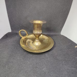 May include: A vintage brass candlestick holder with a circular base and a small handle. The candle holder has a cup-shaped top to hold a candle. The brass has a warm, golden color.