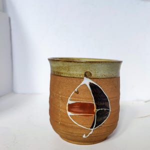 May include: A handmade ceramic mug with a textured, brown exterior and a light green rim. The mug features a painted abstract design in white, black, and red. The mug has a small hole near the rim.