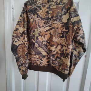 K&ouml;nnte beinhalten: Eine Camouflage-Bomberjacke mit braunem Kragen, Manschetten und Saum. Die Jacke hat ein Muster aus braunen, beigen und gr&uuml;nen Bl&auml;ttern und &Auml;sten. Die Jacke h&auml;ngt an einer wei&szlig;en T&uuml;r.
