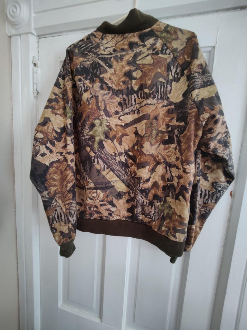 K&ouml;nnte beinhalten: Eine Camouflage-Bomberjacke mit braunem Kragen, Manschetten und Saum. Die Jacke hat ein Muster aus braunen, beigen und gr&uuml;nen Bl&auml;ttern und &Auml;sten. Die Jacke h&auml;ngt an einer wei&szlig;en T&uuml;r.