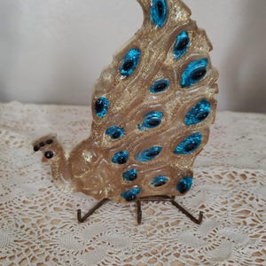 May include: A decorative peacock figurine made of translucent material with gold glitter accents and teal blue eye details. The peacock is mounted on a metal stand, creating a unique home decor item.