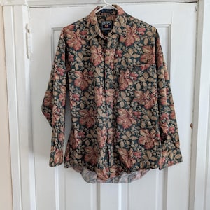 May include: A long-sleeve button-down shirt by Chaps Ralph Lauren featuring a dark green background with a dense, repeating pattern of autumn-colored leaves and berries in shades of rust, orange, and tan. The shirt includes a collar and a chest pocket.