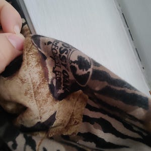 K&ouml;nnte beinhalten: Nahaufnahme eines Stoffabschnitts mit einem Tarnmuster in Braun-, Beige- und Schwarzt&ouml;nen. Der Stoff hat ein Logo mit der Aufschrift "Mossy Oak Forest Floor".