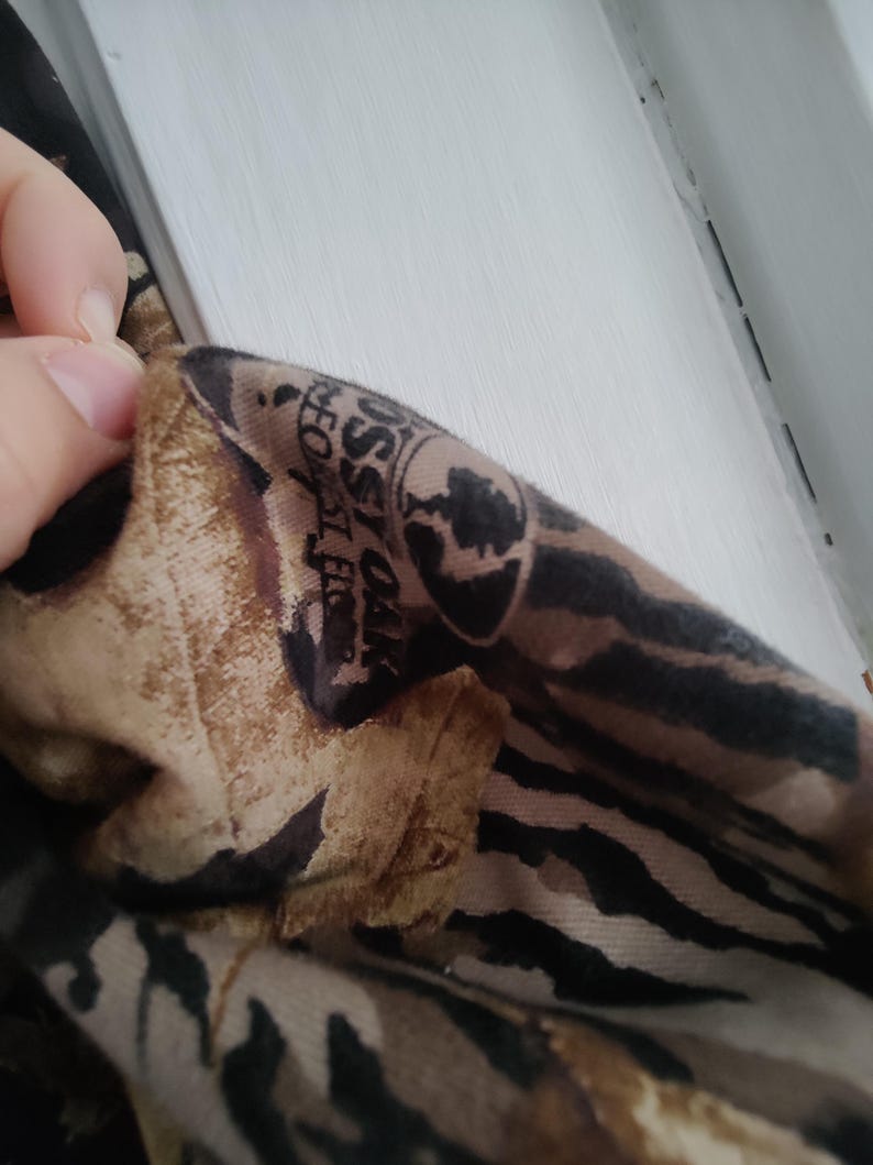 K&ouml;nnte beinhalten: Nahaufnahme eines Stoffabschnitts mit einem Tarnmuster in Braun-, Beige- und Schwarzt&ouml;nen. Der Stoff hat ein Logo mit der Aufschrift "Mossy Oak Forest Floor".