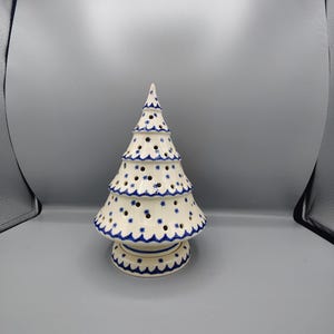 May include: A cream-colored ceramic Christmas tree figurine featuring a hand-painted blue star pattern and scalloped trim. The tree has numerous small circular cutouts throughout its structure and sits on a circular base with a blue wavy border.