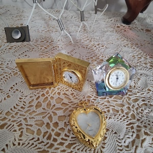 May include: A collection of decorative clocks in various shapes and styles. Includes a gold-colored, heart-shaped clock, a gold-colored rectangular clock, and a crystal-like clock. The clocks have white faces and gold-colored hands.