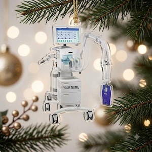 May include: A clear acrylic Christmas tree ornament shaped like a medical ventilator machine. The device features a digital monitor screen, white housing, tubing, and a small purple bag. The text 'YOUR NAME' is printed on the front of the unit.