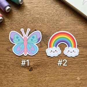 May include: Two die-cut stickers labeled #1 and #2. Sticker #1 is a butterfly with a pink body and light blue and purple wings. Sticker #2 is a colorful rainbow arching over two white clouds, each featuring a simple black face with two eyes and a smile.