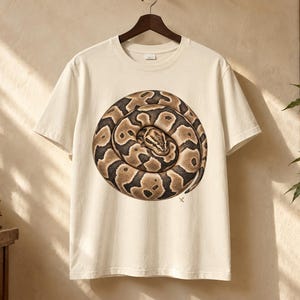May include: A cream-coloured short-sleeve t-shirt featuring a large, circular graphic of a coiled brown and black ball python snake. The snake is depicted in a realistic, detailed style, positioned centrally on the chest of the garment.